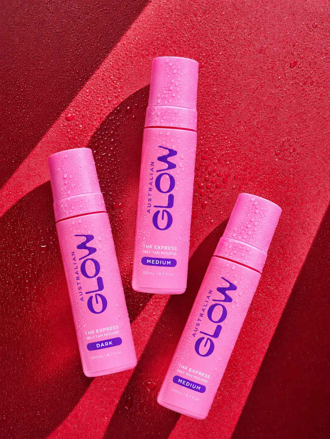 Australian Glow 1HR Express Self Tan Mousse, three pink bottles on red wet surface