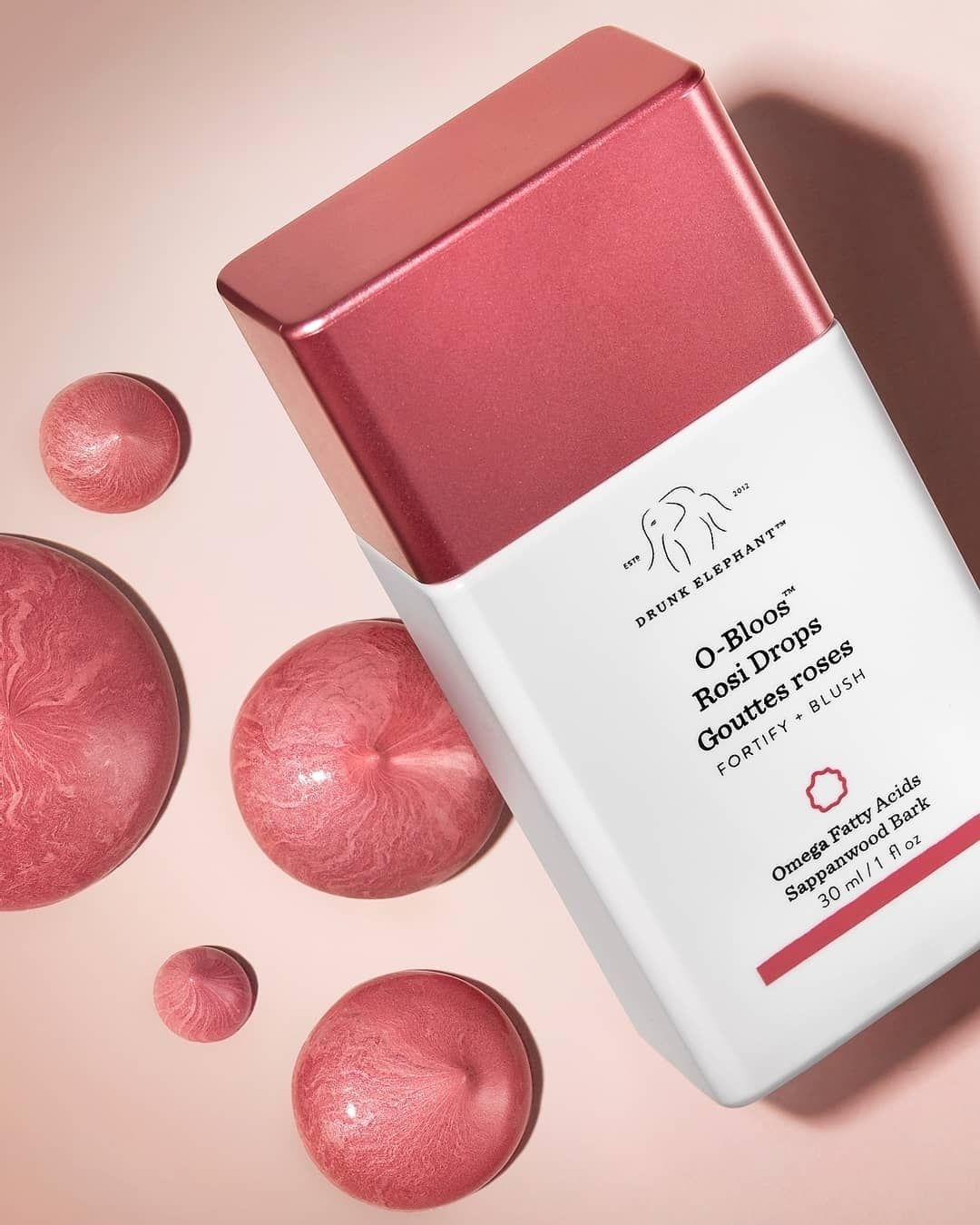 Drunk Elephant O-Bloos Rosi Drops fortifying blush on dusty rose, rose-gold bottle and oil droplets, editorial portrait